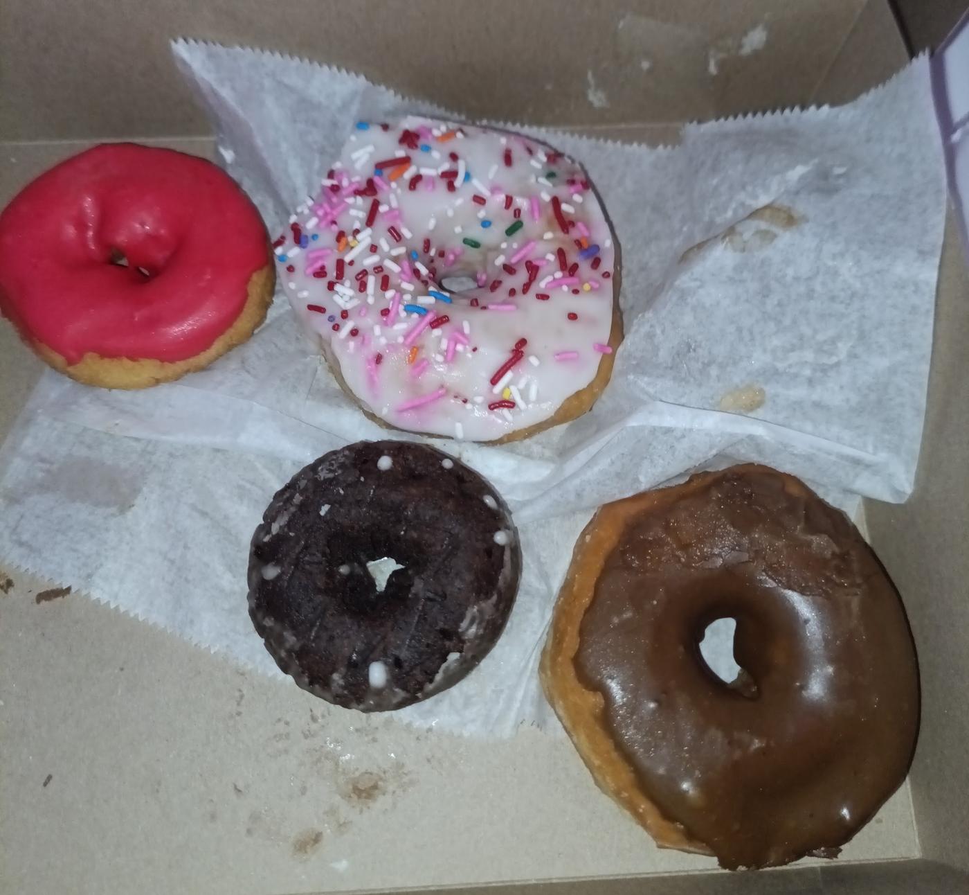 Four donuts in an open kraft box on wax paper: a strawberry iced cake donut, a white iced donut under mixed sprinkles, a chocolate cake donut and a chocolate iced raised donut.